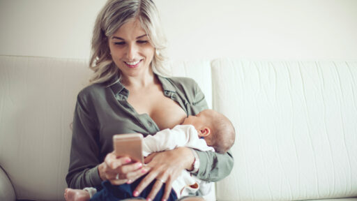 A young White mother breastfeeds while reading on her phone.