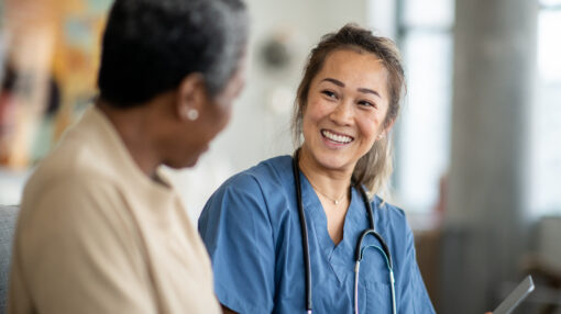 A female nurse of Asian decent, sits with a senior patient in the comfort of her own living room during a Homecare visit. She is wearing blue scrubs and has a tablet in her hands as she reviews some recent test results with her patient. The senior woman is dressed casually and is asking the nurse a question about the results.