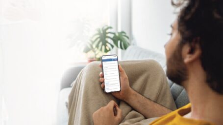 Man in a yellow shirt lounging on a couch, covered with a blanket, looks intently at a smartphone. A plant and soft light add a cozy atmosphere.