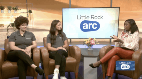 Three women seated in brown leather armchairs during a television studio interview. On the left, a woman with large curly hair wears a gray polo shirt and black pants. In the center, a woman with long straight dark hair wears a matching gray polo shirt and black pants. On the right, a woman in a white patterned blouse and bright orange leather pants gestures with both hands while speaking, holding papers on her lap. A large screen behind them displays the 