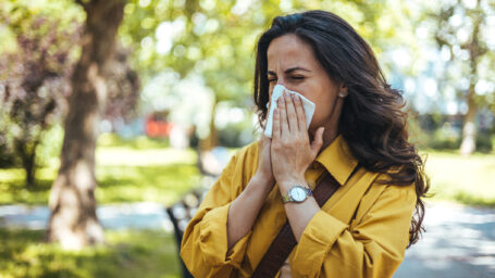 Portrait of unhealthy cute female in yellow top with napkin blowing nose, looks to the source of the allergy, place for advertising. Rhinitis, cold, allergy concept. Pollen allergy symptoms