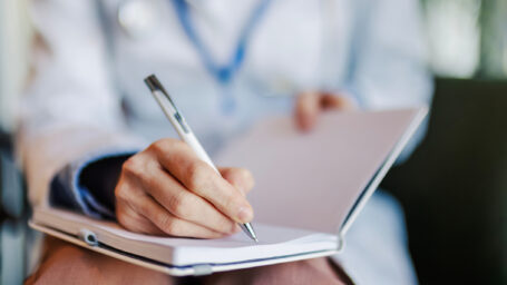 Doctor's hand writing medical notes or a prescription in a notebook, creating a record of patient information and healthcare details during a consultation