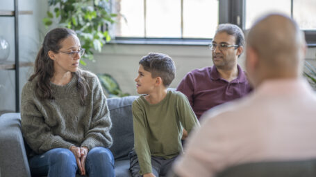 A family of three sit on a couch in front of a counselor and discuss mental health struggles in an urban office space